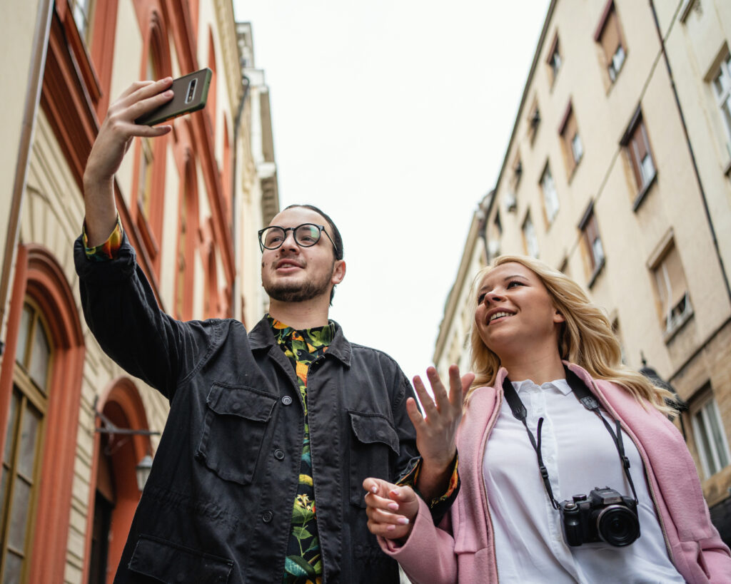 Two people walking in the street with holding the camera up to record themselves and one having a camera around their neck